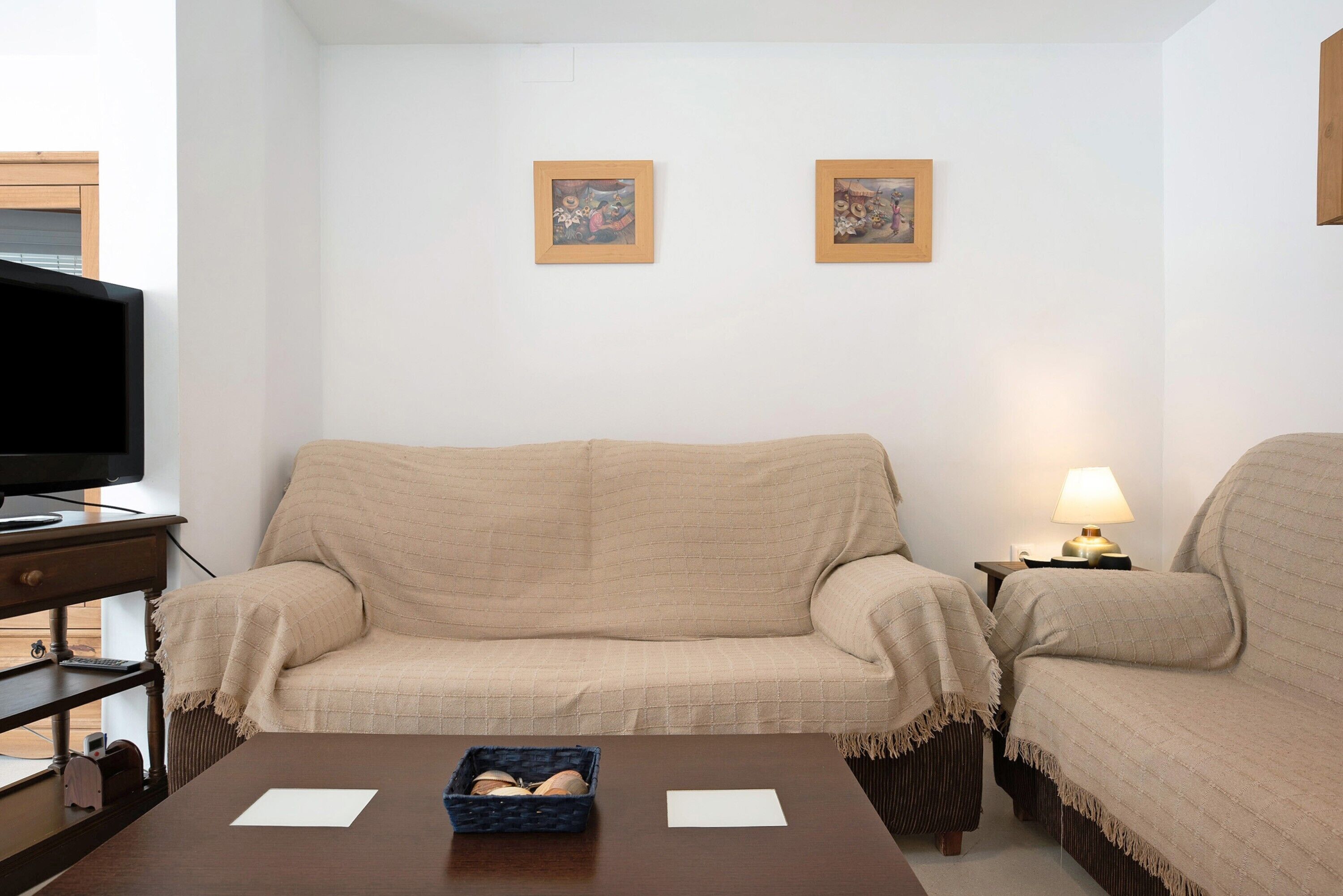 Een woonkamer met een beige bank, een televisie op een meubel, een kleine tafel met een mand en papieren, en twee ingelijste foto's aan de muur.
