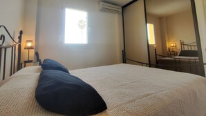A bedroom with a bed, a window, an air conditioner, a mirror, and a nightstand with a lamp.