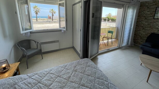 A room with a bed, a chair, a table, and a balcony with a view of the beach.