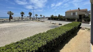 A coastal road with a sandy area, palm trees, and a building with a tiled roof.