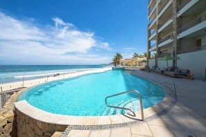 A pool with a curved edge, a stone border, and a view of the beach and ocean.