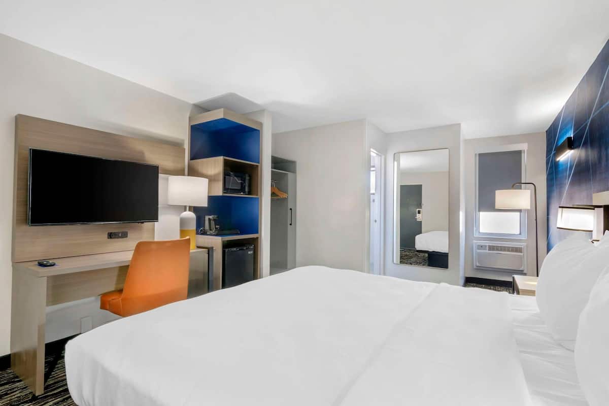Habitación de hotel con cama, televisor, escritorio, silla, armario y espejo.