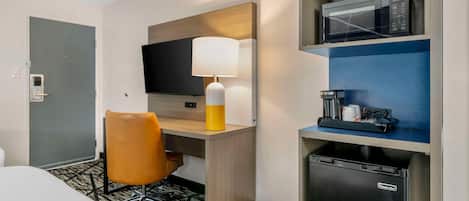 A hotel room with a bed, a desk with a lamp, a television, a microwave, a coffee machine, and a mini-fridge.