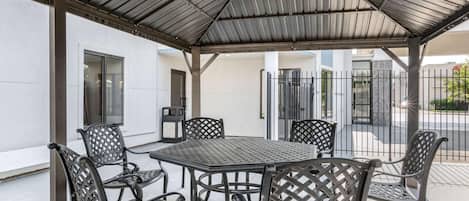 A covered outdoor seating area with a table and chairs, a trash bin, and a building entrance.
