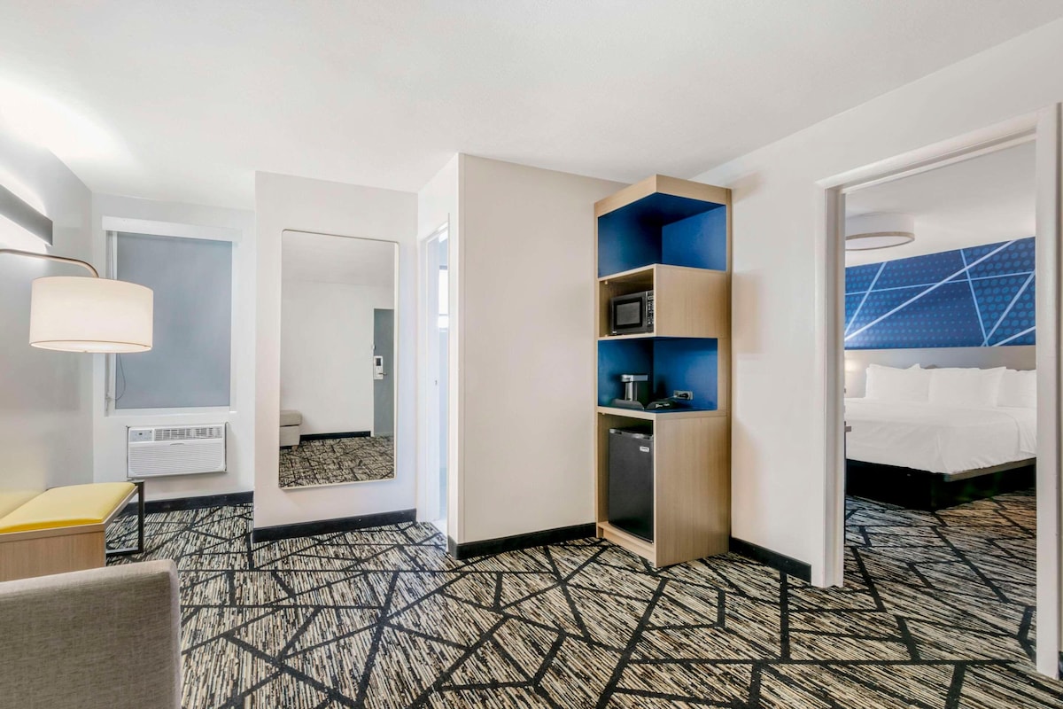 A hotel room with a geometric patterned carpet, a built-in TV shelf, a bedside table, and a bed with a blue headboard.