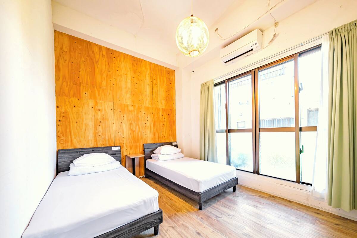 A hotel room with two beds, wooden paneling on one wall, a large window with curtains, and an air conditioning unit.