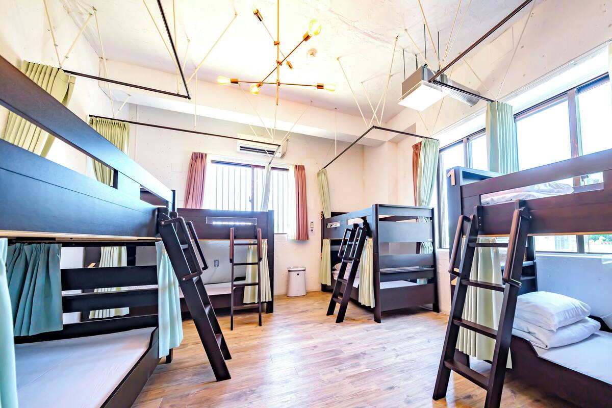 A dormitory room with bunk beds, a ladder, a radiator, and a window with curtains.