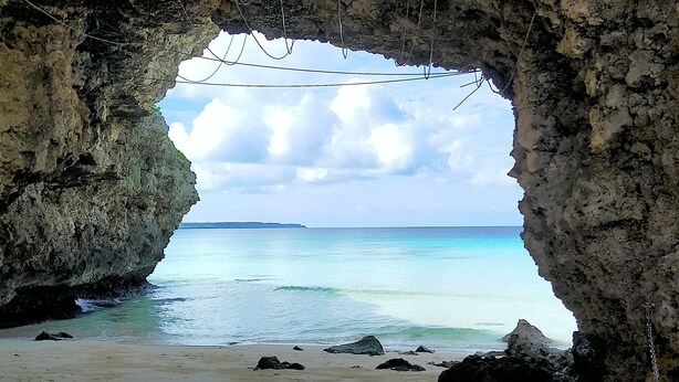 Blick auf einen Strand durch einen Felseneingang, mit klarem türkisblauem Wasser und einem entfernten Landteil.