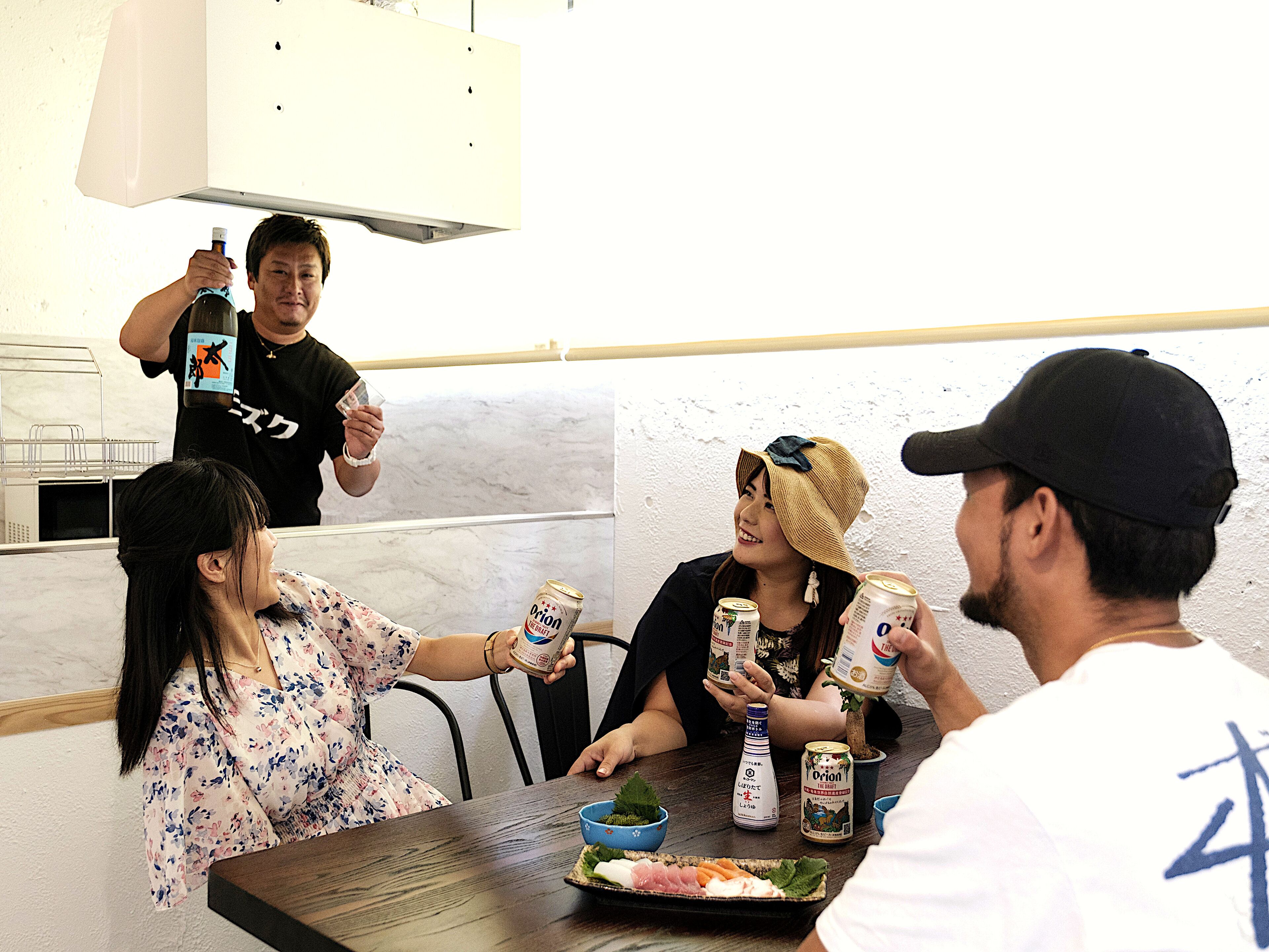 A group of people sitting around a table with food and drinks, including bottles and cans.