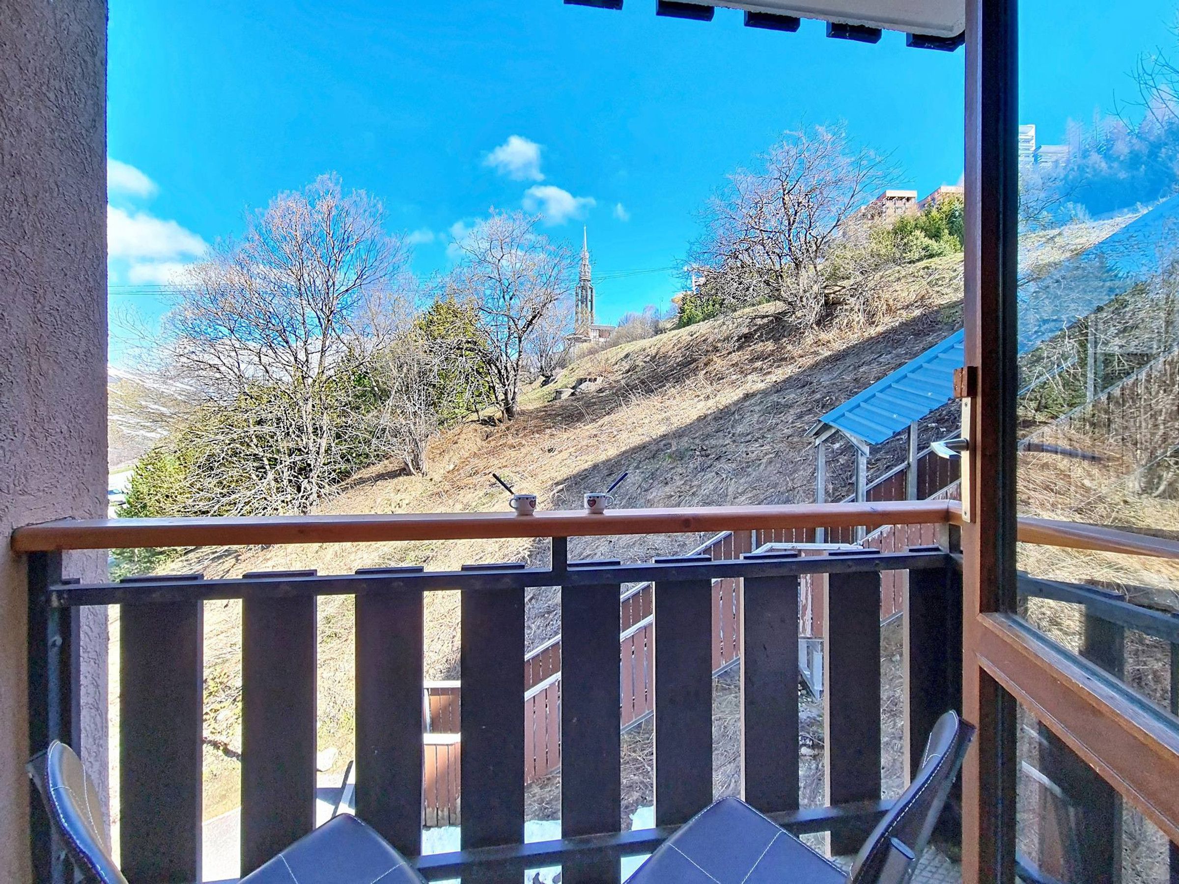 A balcony with a view of a hillside, a wooden railing, and a small table with a cup and saucer.