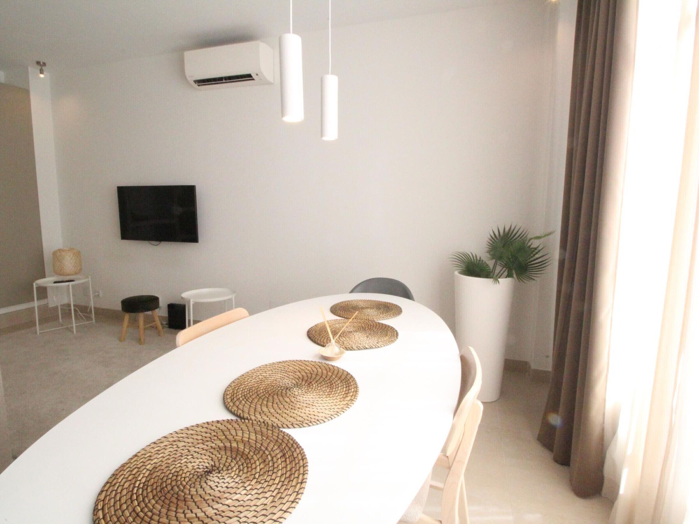Een moderne eetruimte met een witte tafel, geweven placematen, hangende吊灯 en een pot met een plant.
