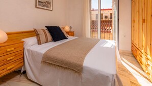 A bedroom with a wooden bed, white bedding, a wooden nightstand, and a view of a tiled roof through a glass door. - Charming Village House with Terrace, Near Beach, WiFi, Garage & Fully Equipped Kitchen (Palamós)