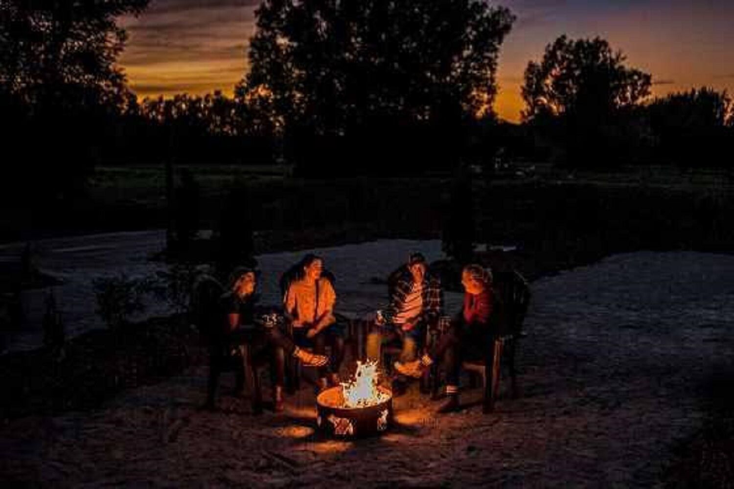 Um grupo de pessoas sentadas ao redor de uma fogueira em um cenário rural ao anoitecer.