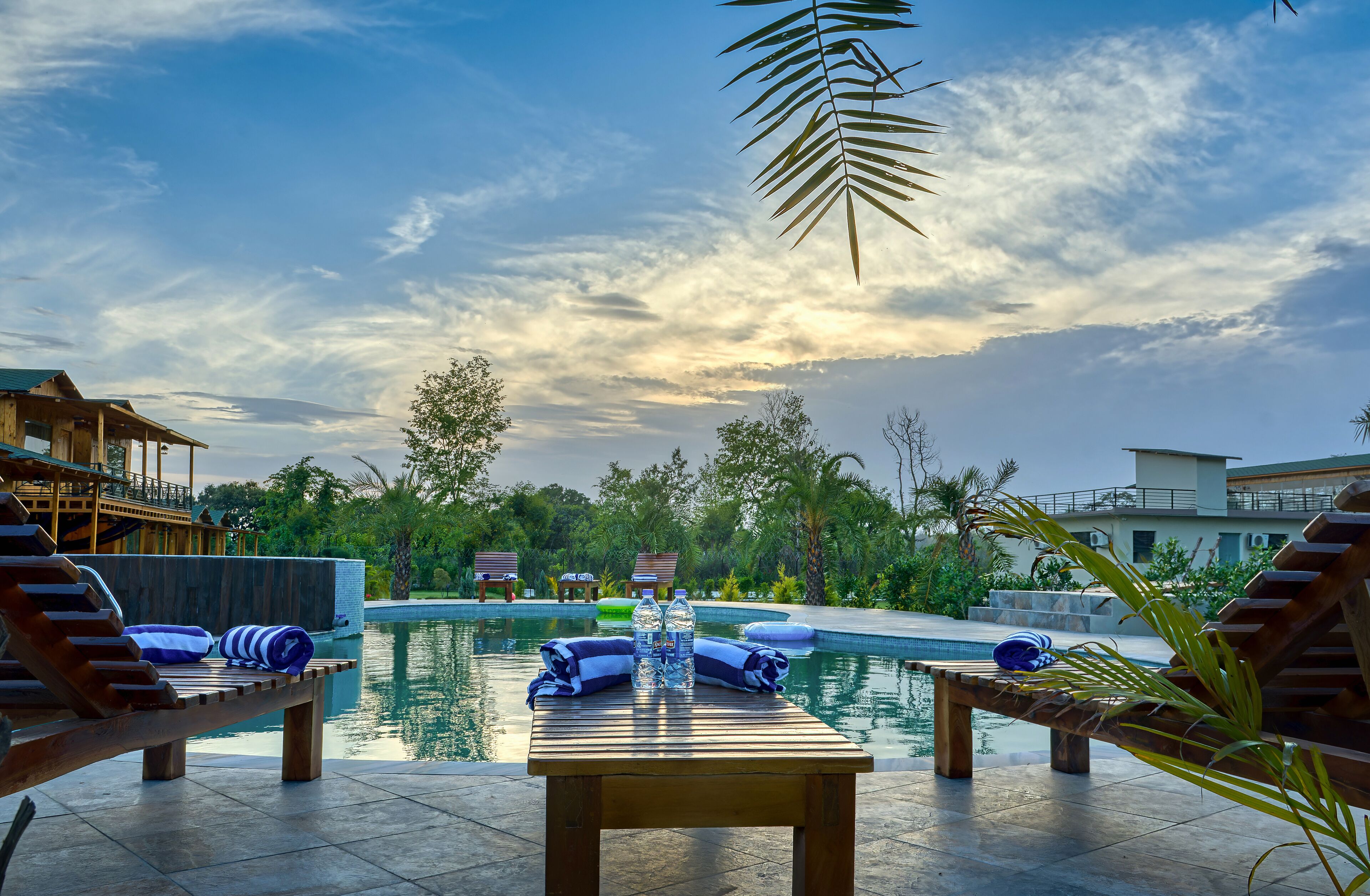 A poolside area with lounge chairs, a wooden table with towels and water bottles, and a view of the pool and surrounding greenery.
