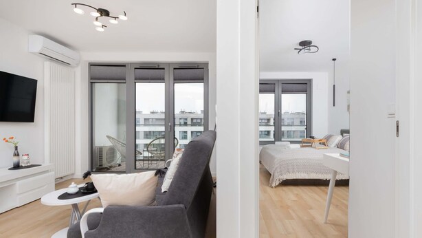 A modern apartment with a grey sofa, a small white table, a TV, and a balcony with a view of buildings.