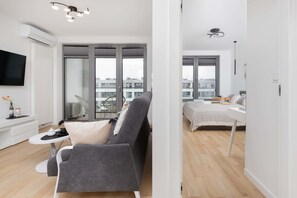 Un appartement moderne avec un canapé gris, une petite table blanche, un téléviseur et un balcon offrant une vue sur les immeubles.