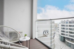 Un balcon avec des fauteuils en osier, une petite table ornée d’une plante et une vue sur des immeubles modernes.