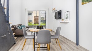 A modern living room with a sofa, dining table, and chairs, a TV mounted on the wall, and a large window with a view of a building and greenery outside. - Venus Apartments - Westside Residence (Budapest)