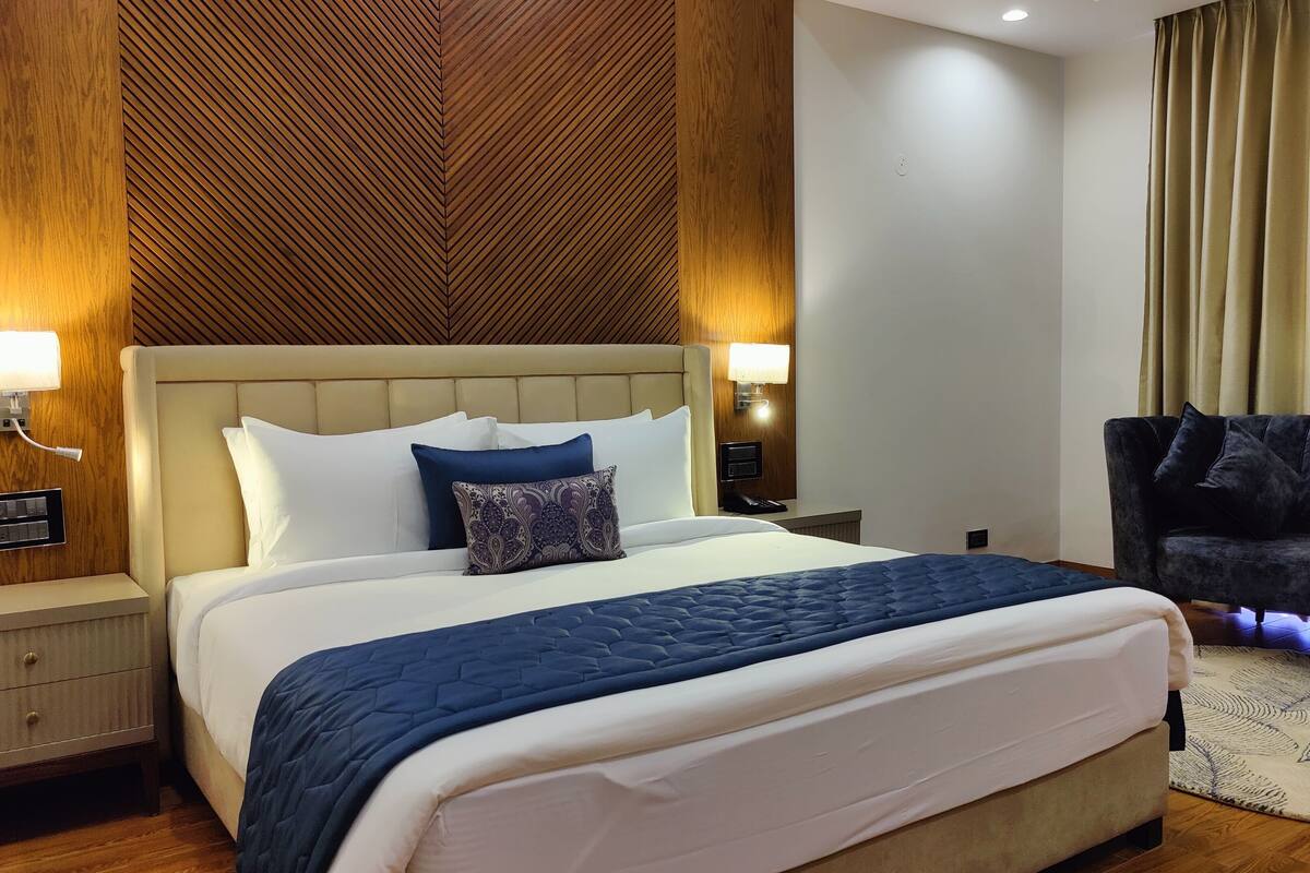 Una habitación de hotel con una cama grande, dos lámparas de noche, un sillón oscuro y una pared de acento de madera.