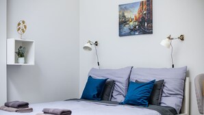 Una cama bien hecha con edredón blanco y gris, almohadas azules y grises, y toallas dobladas a un lado. Hay un estante de pared con una planta en maceta y un adorno metálico. Un cuadro de un paisaje urbano cuelga sobre la cama.