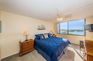 A bedroom with a bed, nightstands, a ceiling fan, a large window with a view, and a TV on the wall.
