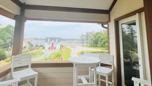 A covered porch with two white chairs and a small table, overlooking a house and a body of water.
