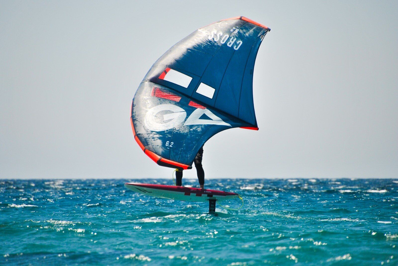 Eine Person windsurft bei klarem Wetter. Das Segel trägt die Aufschrift 'Cross 62'.