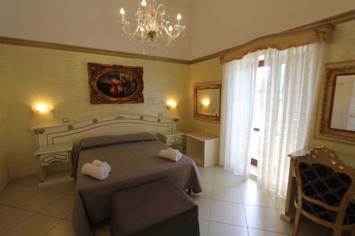 A hotel room with a large bed, a chair, a desk, a chandelier, a painting, and a mirror.