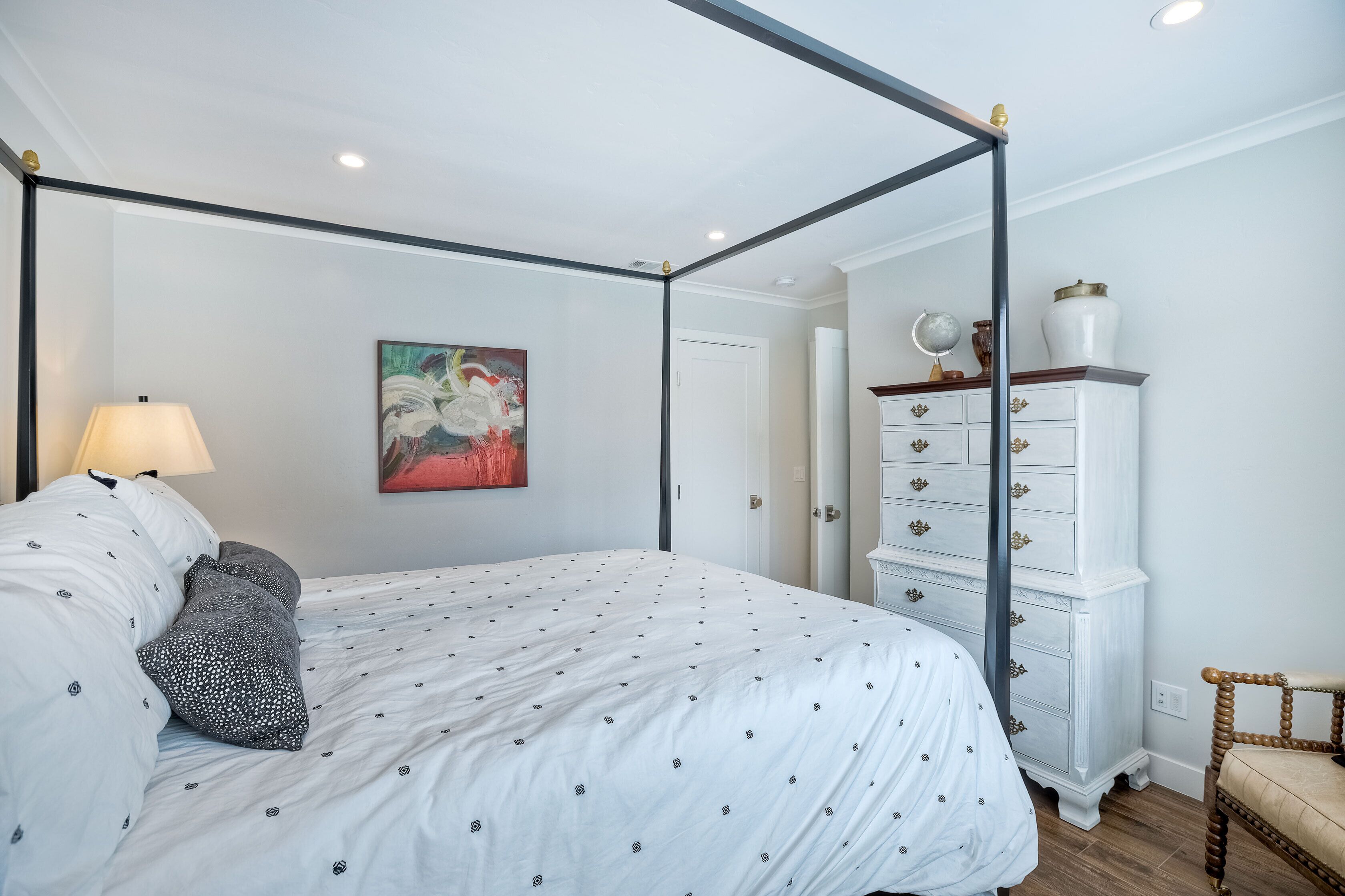 Un lit à baldaquin avec un couvre-lit blanc et des oreillers gris, une commode blanche avec des poignées dorées et une chaise en bois.