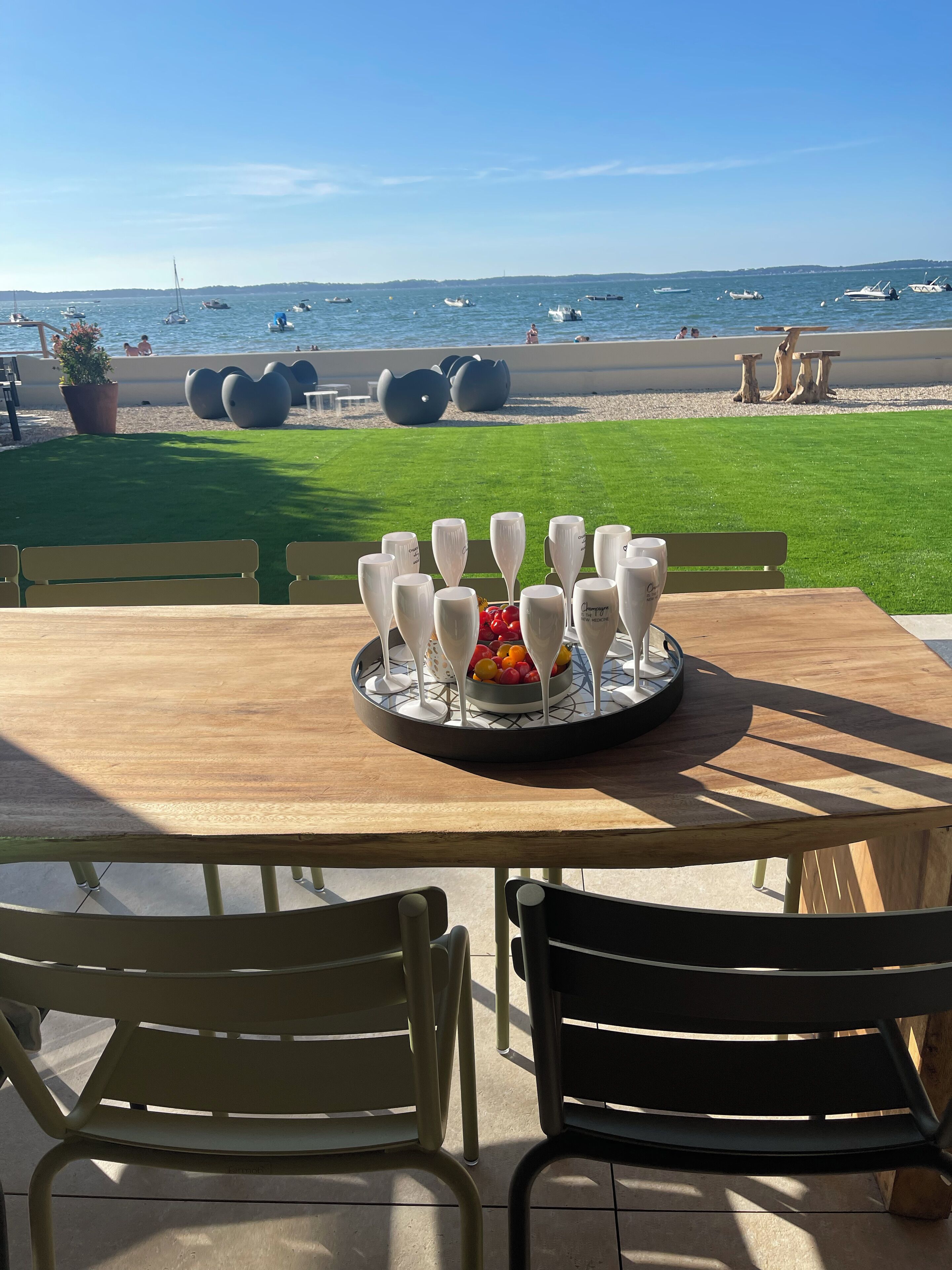Ein Tisch mit Obstdekoration und weißen Gläsern, mit Blick auf einen Strand mit Booten und Menschen.