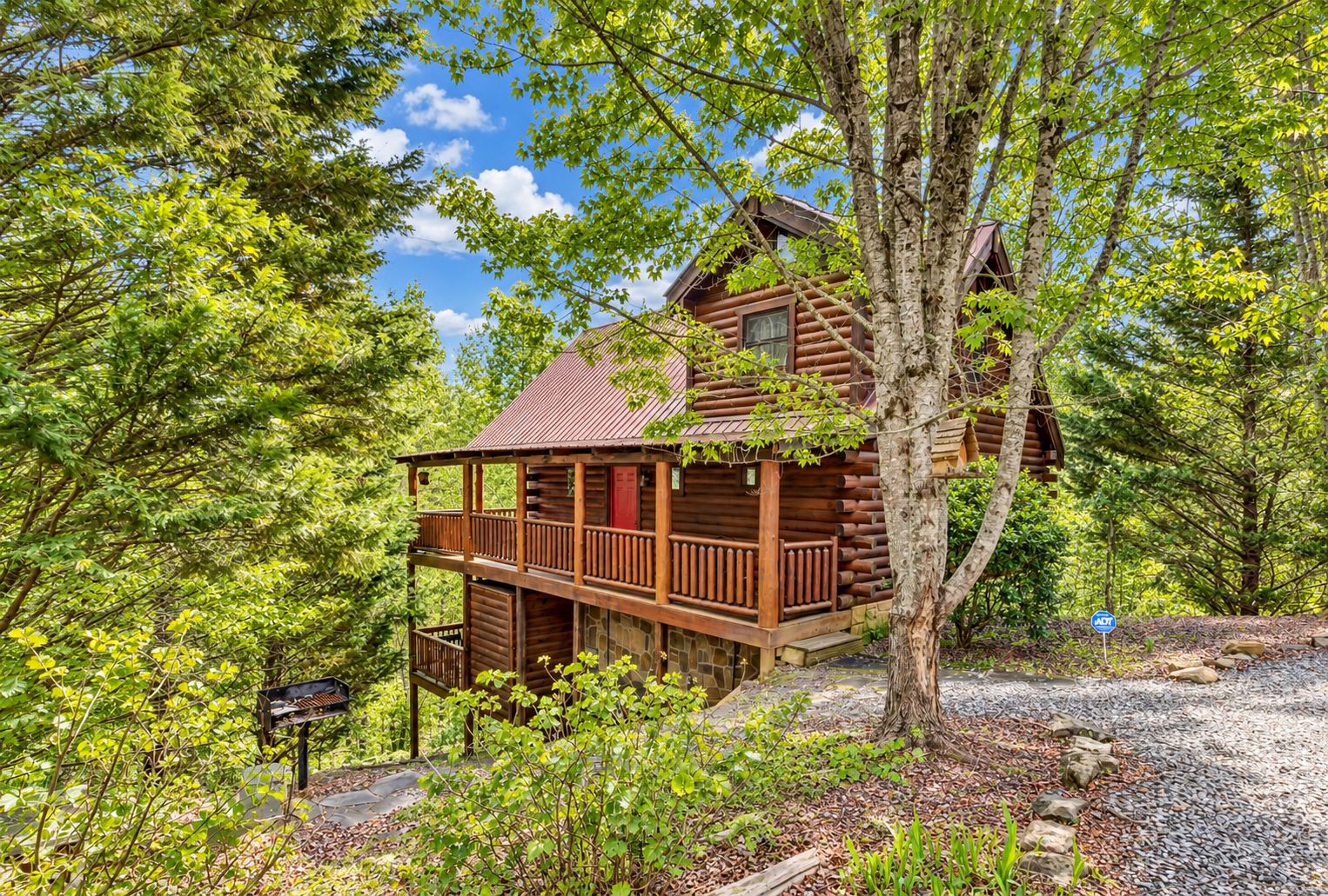 A two-story log cabin with a balcony, surrounded by dense trees and a gravel path.