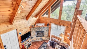 Interior acogedor de una cabaña con chimenea de piedra, vigas de madera, televisor y ventana con vistas a la vegetación.