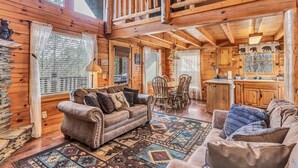Interior acogedor de una cabaña de troncos con comedor de madera, cocina con armarios de madera y sala de estar con sofá y alfombra estampada.