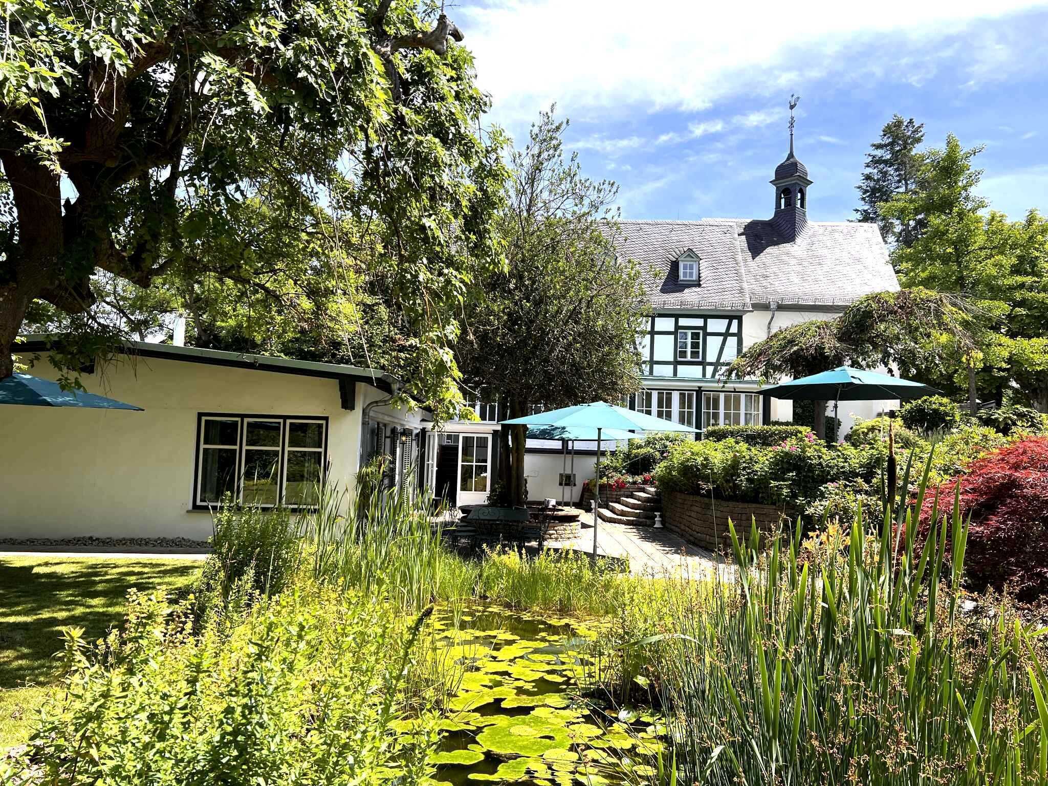 A traditional Tudor-style building with a prominent tower, surrounded by lush gardens and a pond with lily pads.