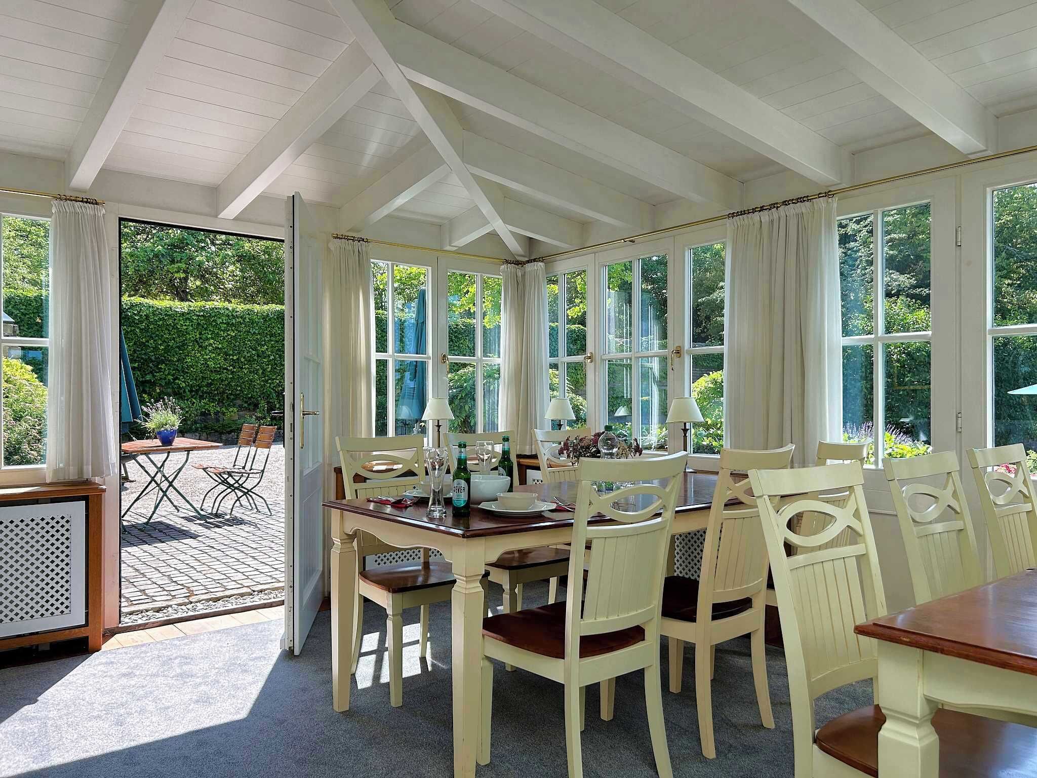 A dining area with a table set for six, surrounded by white chairs, under a white ceiling with exposed beams.
