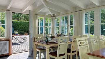 A dining area with a table set for six, surrounded by white chairs, under a white ceiling with exposed beams.