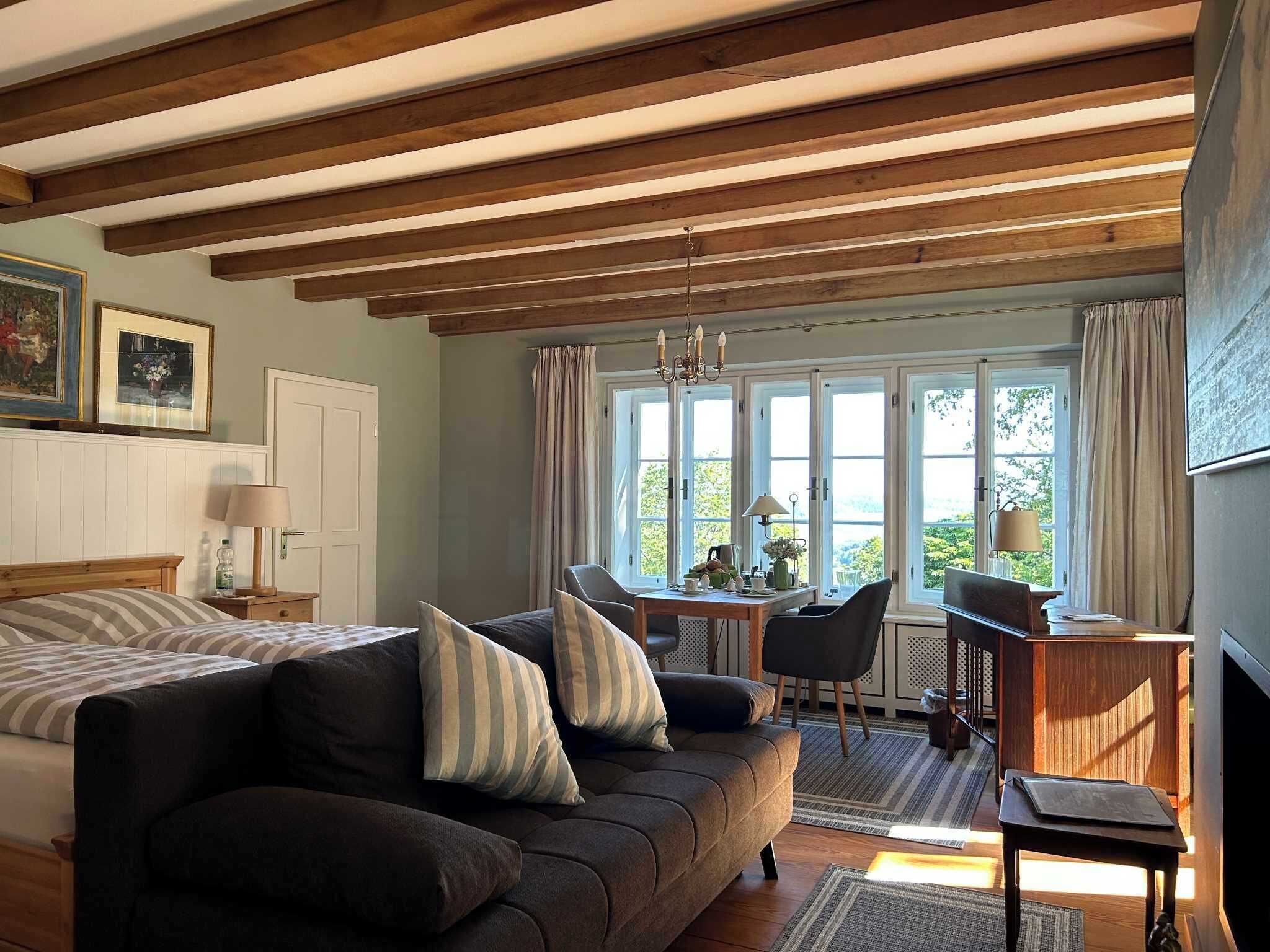 A cozy room with a bed, a sofa, a dining table, and a TV. There are windows with curtains, a chandelier, and a wooden ceiling.