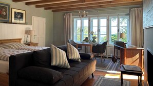 Um quarto aconchegante com cama, sofá, mesa de jantar e televisão. Há janelas com cortinas, um lustre e teto de madeira.