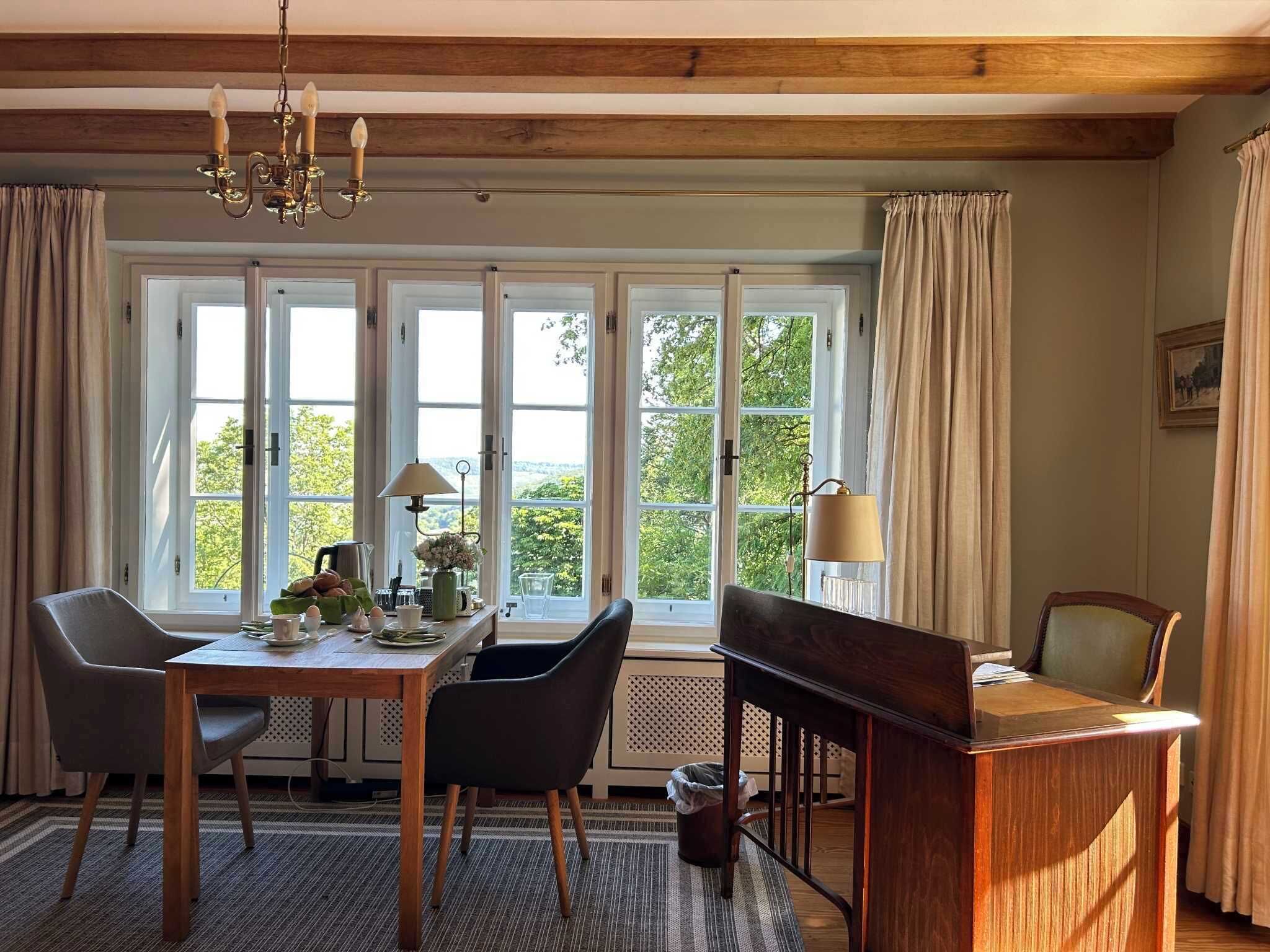 A room with a wooden table, two chairs, a desk, and a window with curtains.
