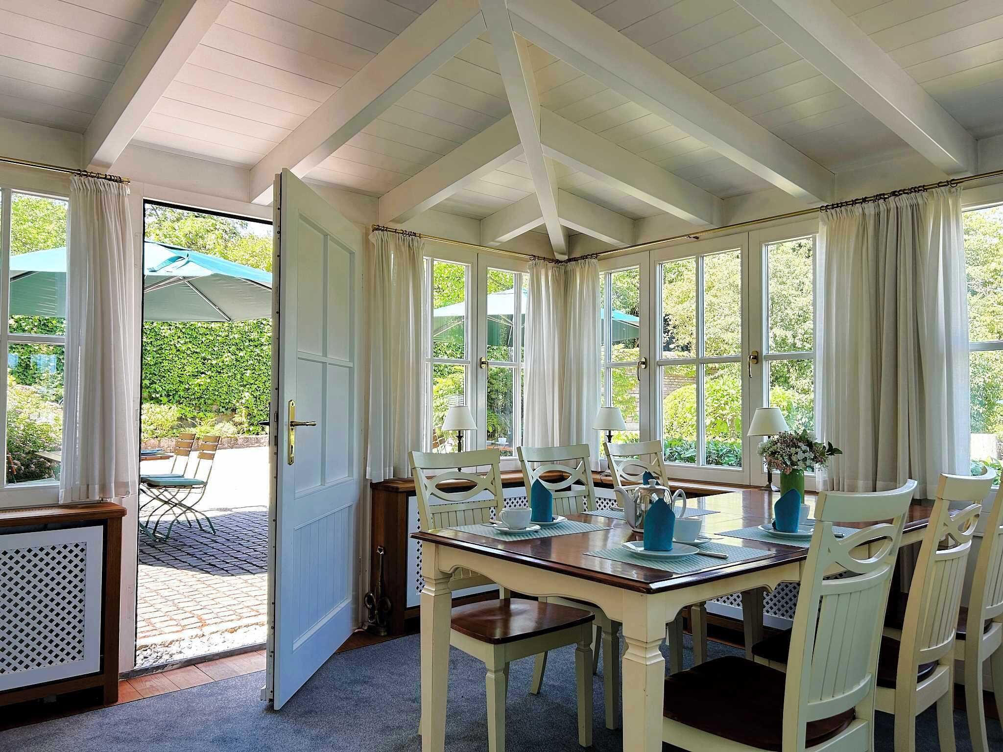 A dining area with a wooden table and chairs, large windows with curtains, and a view of greenery outside.