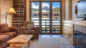 A cozy living room with a leather sofa, a wooden coffee table, a plaid curtain, and a view of a mountain and a building through large glass doors. - Summit View- Big Sky Shoshone Ski in/Out, Heated Pool (Big Sky)