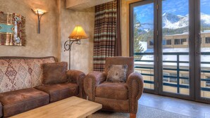 A cozy room with a leather sofa, armchair, and wooden table. It features a large window with a view of mountains and a balcony. - Summit View- Big Sky Shoshone Ski in/Out, Heated Pool (Big Sky)