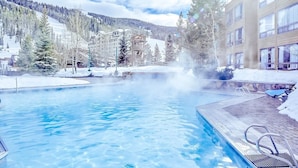 Ein Pool mit klarem Wasser, umgeben von schneebedecktem Boden und Bäumen, mit einem Gebäude im Hintergrund.