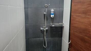 Un petit espace douche fermé, avec des carreaux de couleur sombre, une pomme de douche à main, un distributeur de savon fixé au mur et une porte en bois.