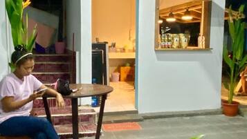 Una persona sentada en un banco en una habitación con una pequeña mesa, una ventana con un estante y plantas en macetas.