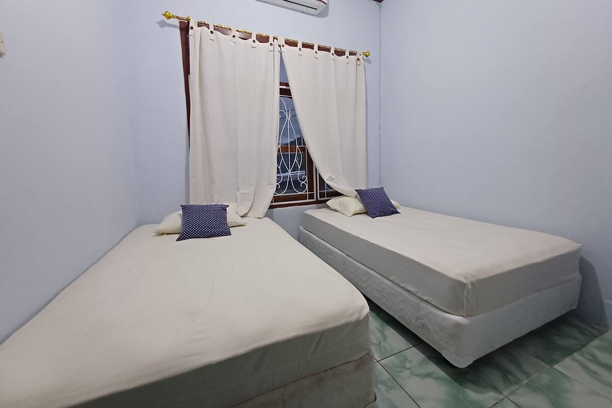 A small, clean room with two single beds, a window with curtains, an air conditioning unit, and a tiled floor.