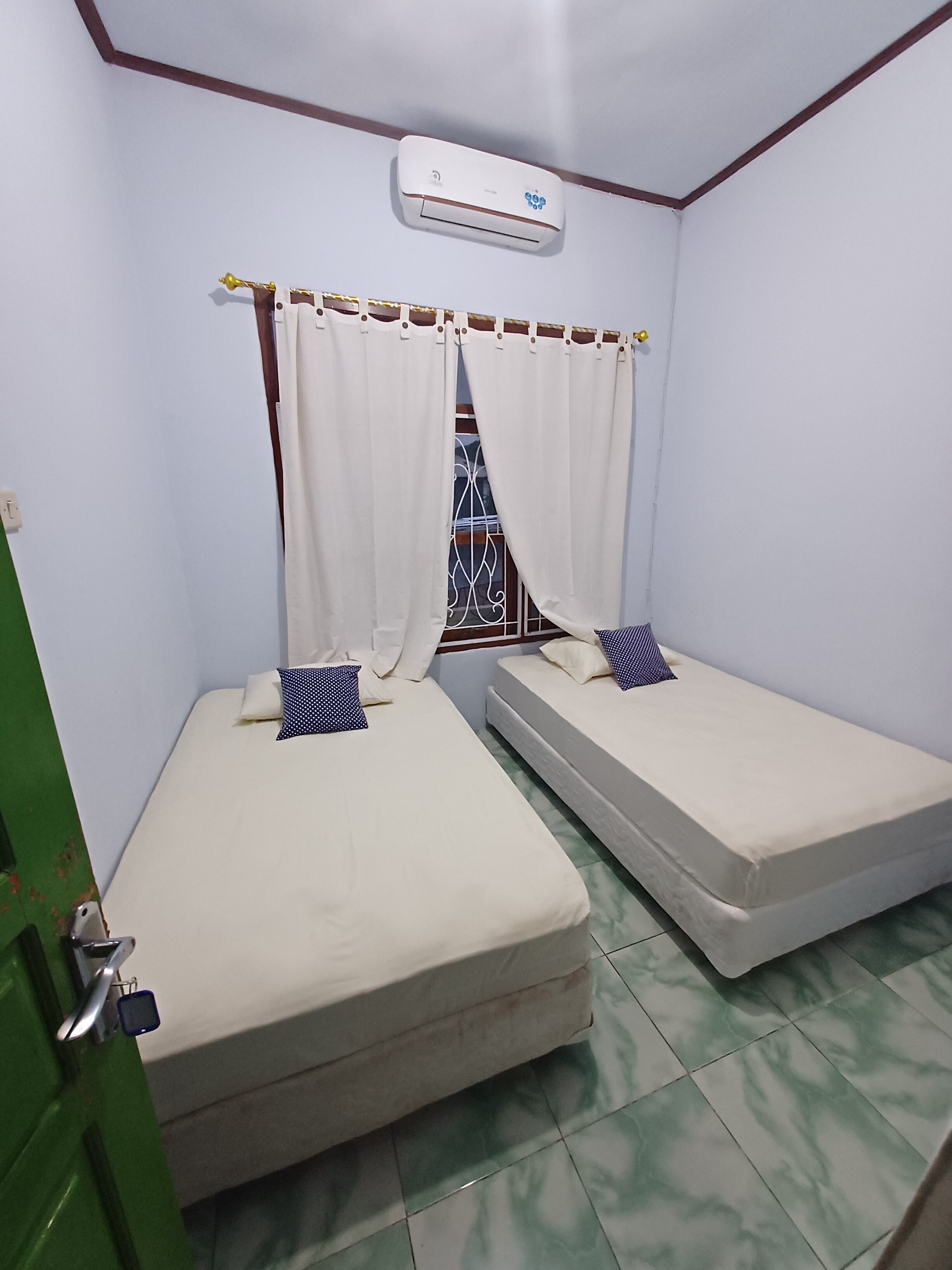 A small, single-room accommodation with two beds, a window with metal bars, a white air conditioning unit, and a green door.