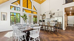 A spacious dining area with a wooden table and white chairs, large windows, and a view of the outdoors.