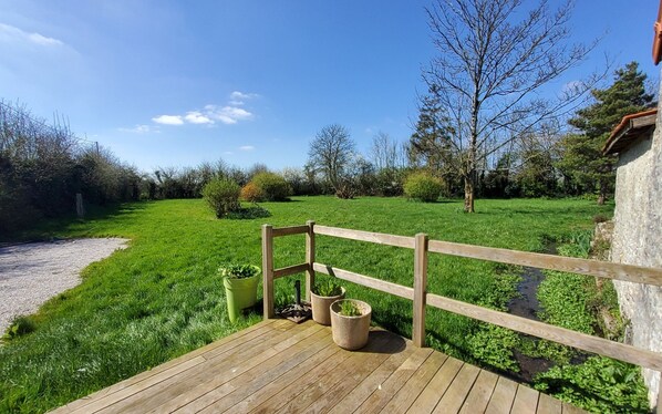 A wooden deck with potted plants, a stone wall, and a grassy field with trees. - Charming, renovated barn on the outskirts of Sainte-Mère-Église (Sainte-Mère-Église)