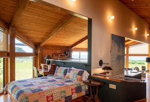 A cozy room with a wooden ceiling, a large bed with a colorful quilt, a desk with a lamp, and a view of the outdoors. - Tin Roof | Courtyard Retreat Near Bluff Trails (Sea Ranch)